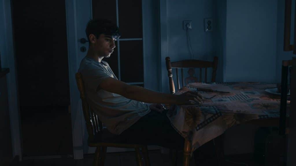 A young man sitting at a table in a dark room looking away from the camera.