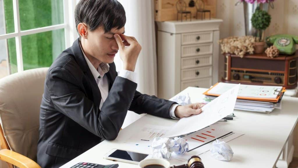 A man in a suit sits at a desk holding papers while pinching his nose bridge.