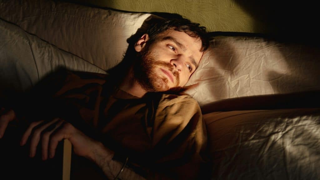 A man with a beard lies in bed looking sideways in low, dramatic morning light.