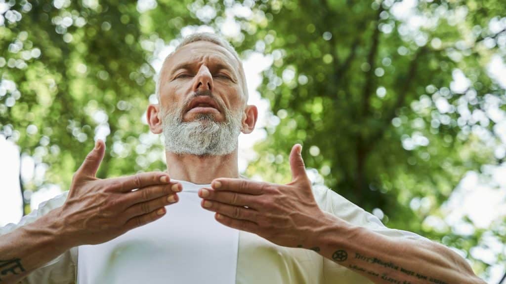 A man with a grey beard stands outside with closed eyes and his hands raised.