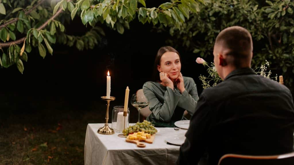 A woman sits at a candlelit table outdoors, looking at a man from across the table.