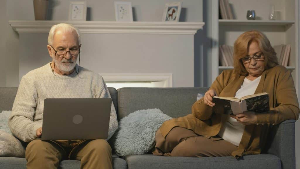 A man uses a laptop while a woman reads a book on a grey sofa.