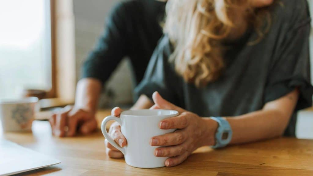 A person holding a white mug while sitting at a wooden table.