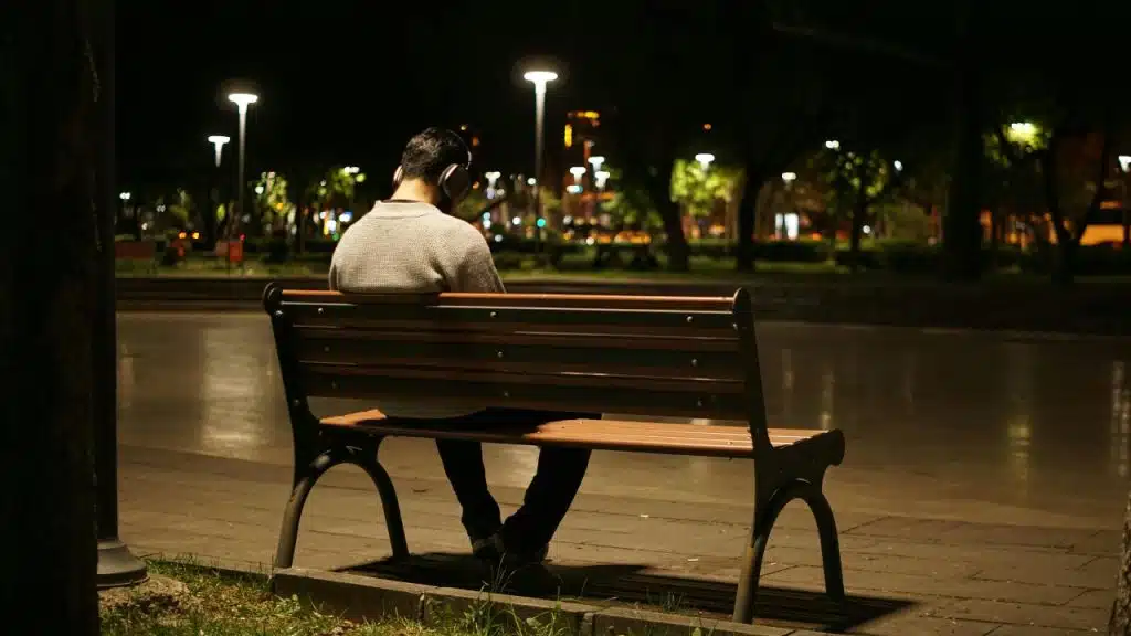 A person wearing headphones sits alone on a wooden park bench at night under street lights.