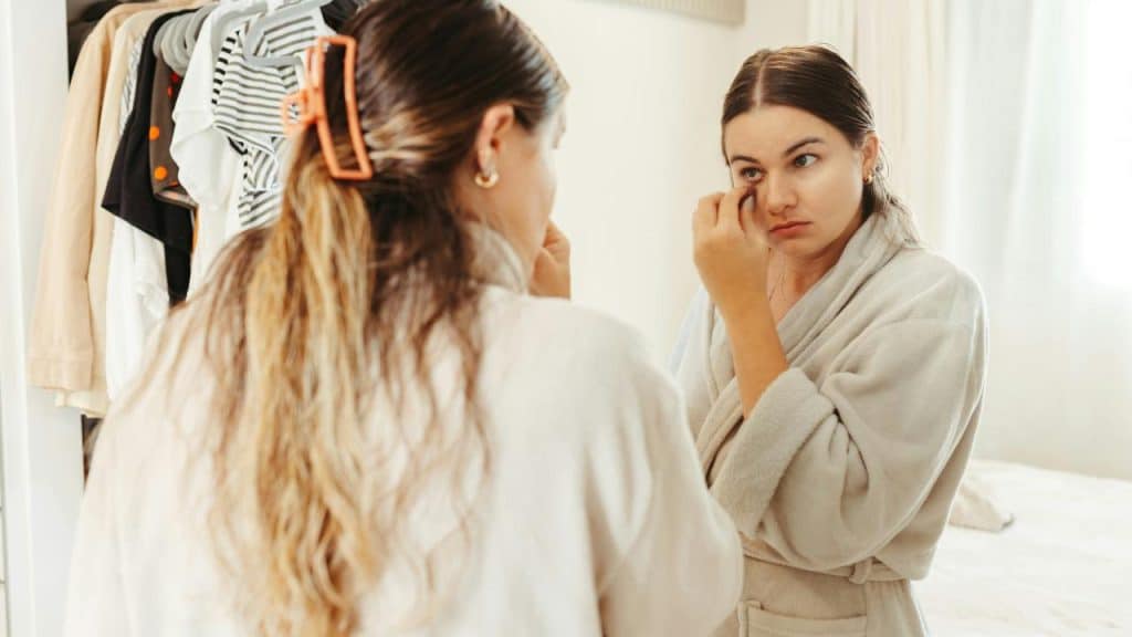 A woman in a robe looking at herself in a mirror while applying makeup.