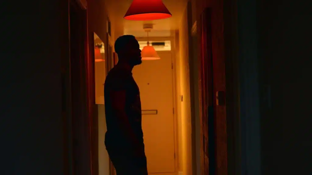 A man stands in a dark hallway illuminated by an orange glow from overhead lamps.