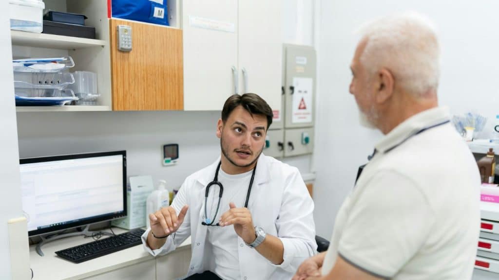 A man in a white lab coat with a stethoscope speaks to a seated patient.
