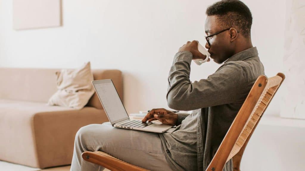 A man sitting on a chair drinking coffee while working on a laptop.