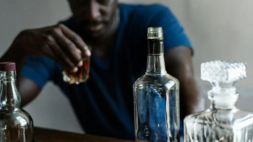 A man reaches for a small glass of liquid behind several clear and brown bottles.
