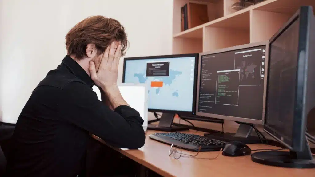 A man in a black shirt covers his face while sitting before three computer monitors.