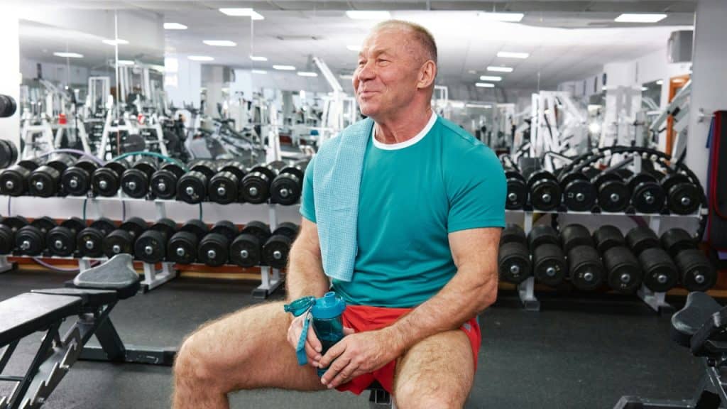A smiling man in gym attire sits on a bench holding a blue water bottle.