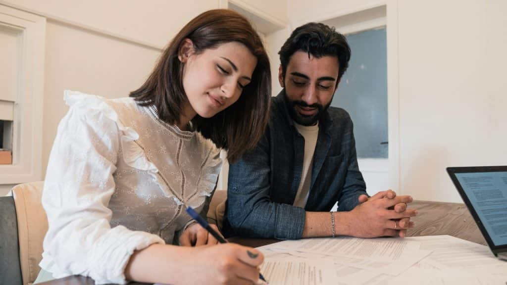 A woman sits at a wooden table signing documents while a man watches her closely.