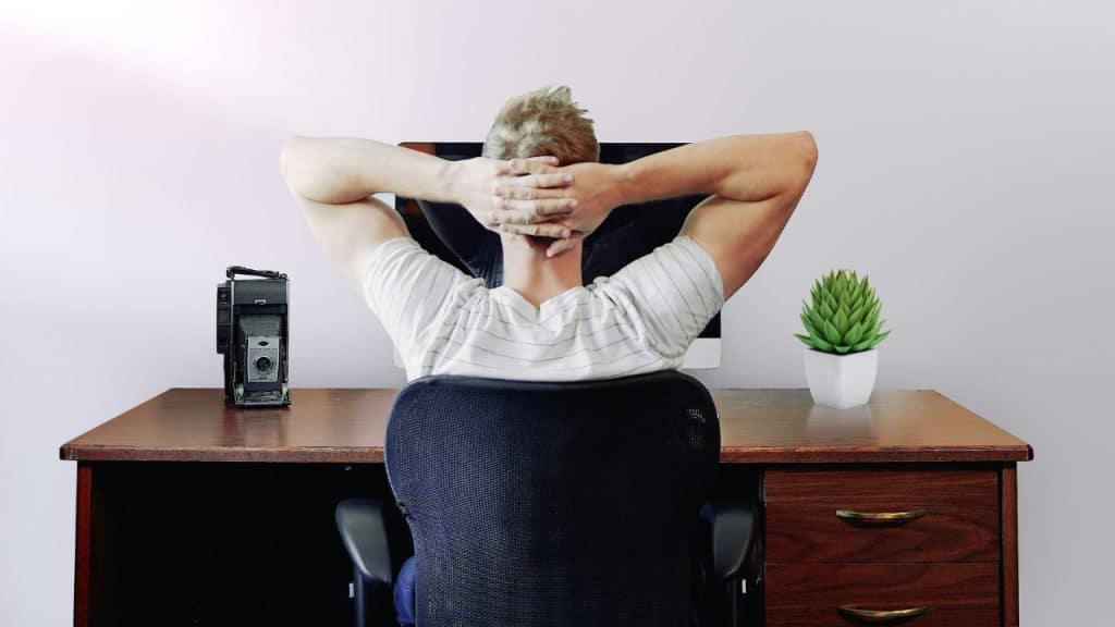 A man sits at a desk with his hands behind his head, facing a computer monitor.