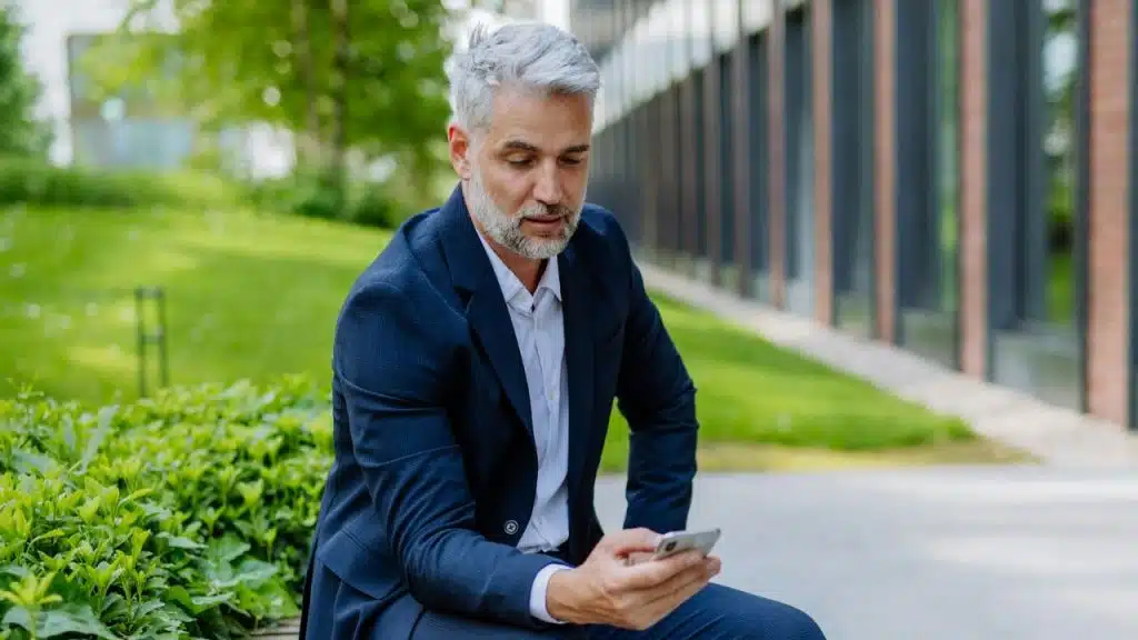 A man with gray hair in a blue suit sits outside while looking at a smartphone.