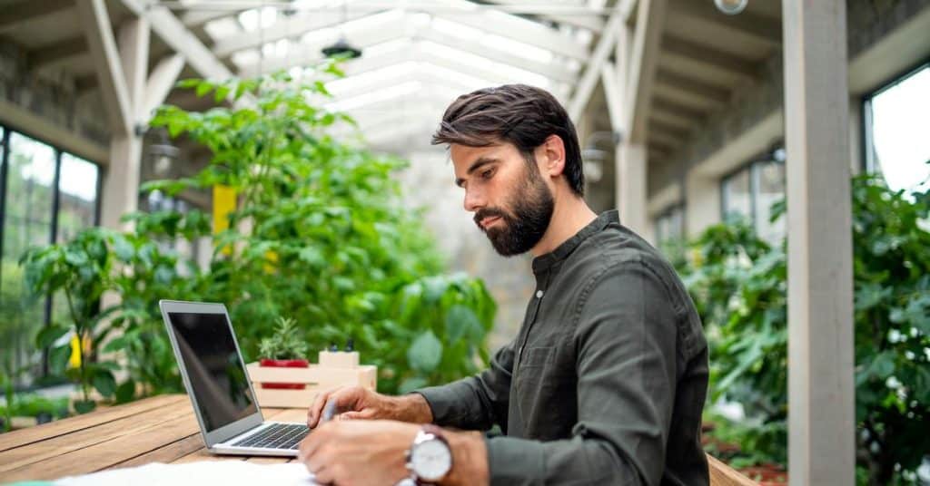 A man works on a laptop at a table surrounded by plants.