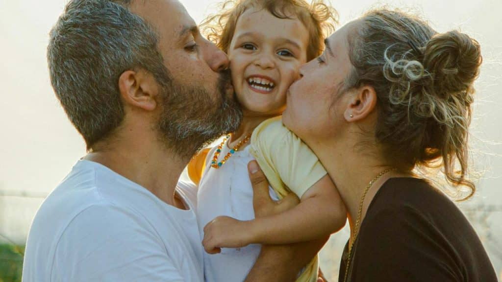 A child smiling as two adults kiss their cheeks.