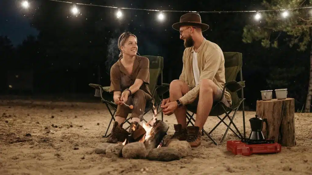 A couple sitting by a campfire at night, talking and roasting food
