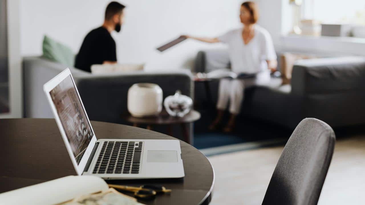 A laptop on a table with two people talking on a couch in the background.