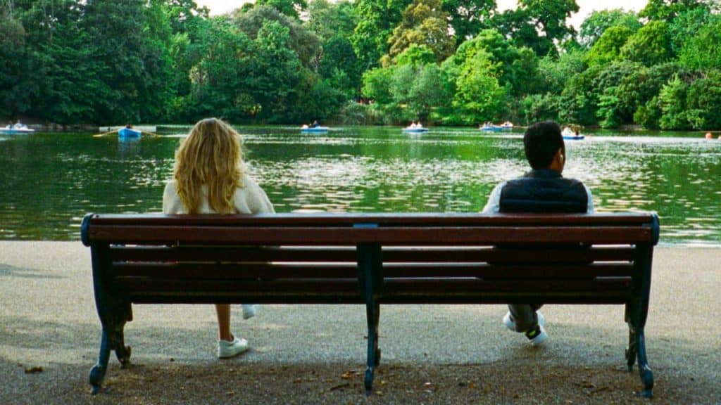 A woman and man sit on opposite ends of a park bench facing a lake.