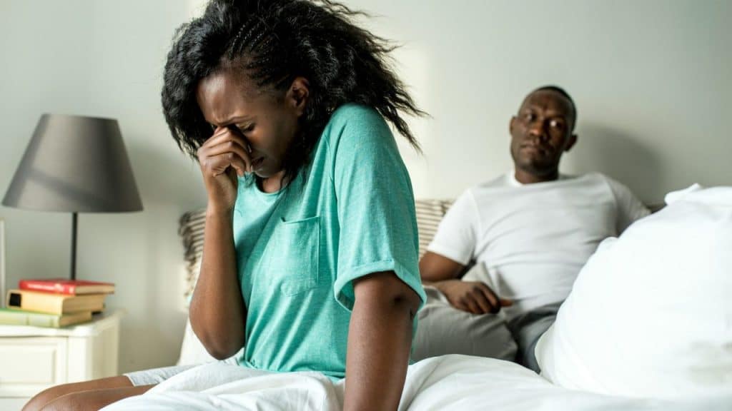 A woman sits on a bed covering her face while a man watches from the background.