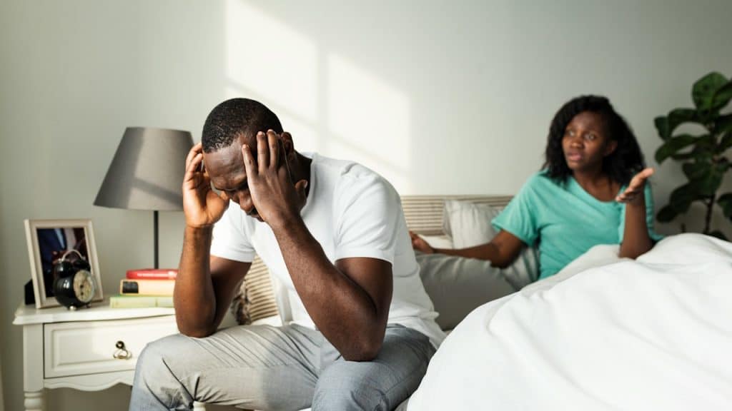 A distressed man sits on the edge of a bed with his head in his hands while a woman gestures argumentatively behind him.