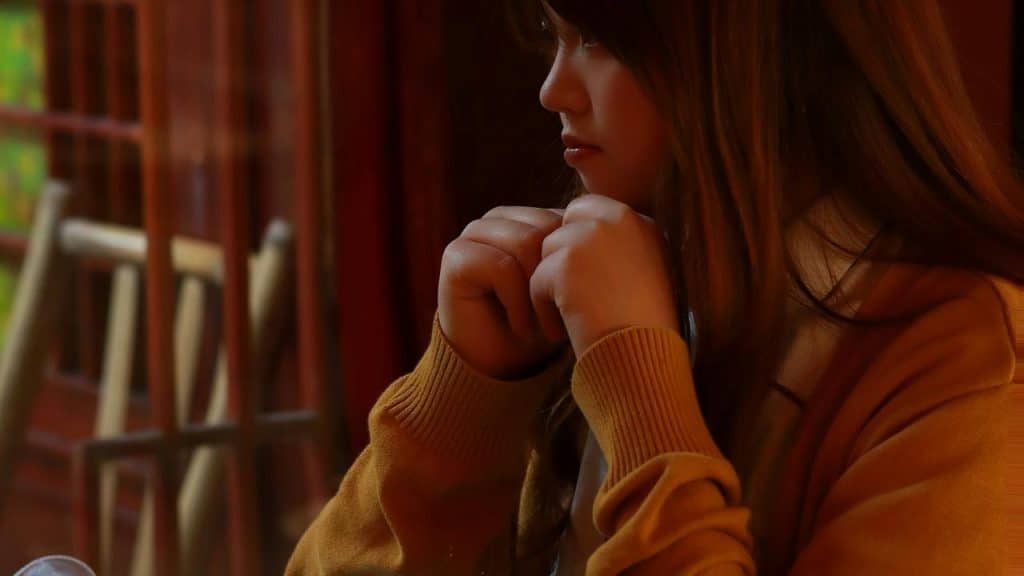 A person sits indoors with hands clasped near their chin, looking thoughtful.