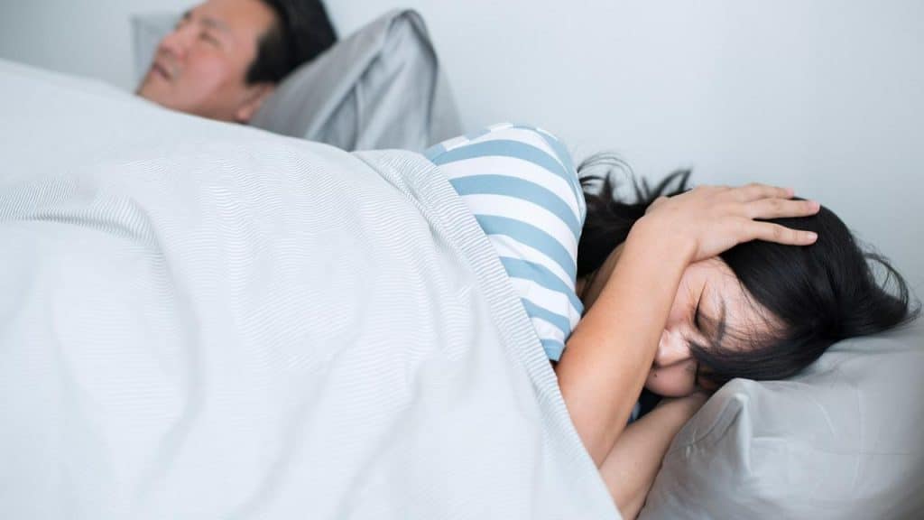 A woman in bed covering her ears while a man sleeps beside her.