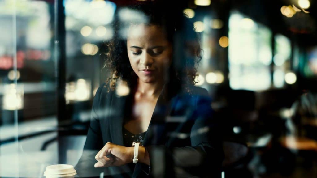 A woman in a black blazer looks down at her wristwatch inside a dimly lit cafe.
