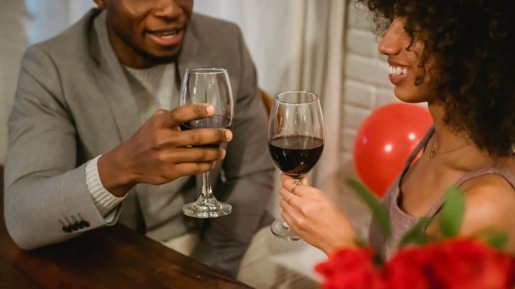 A man and woman hold glasses of red wine while smiling at a wooden table.
