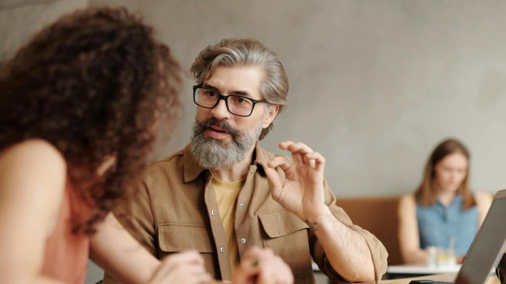 A bearded man with glasses gestures while talking to a woman with curly hair indoors.