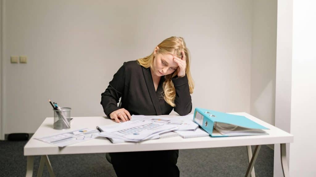 A blonde woman in a black blazer sits at a desk looking at papers.