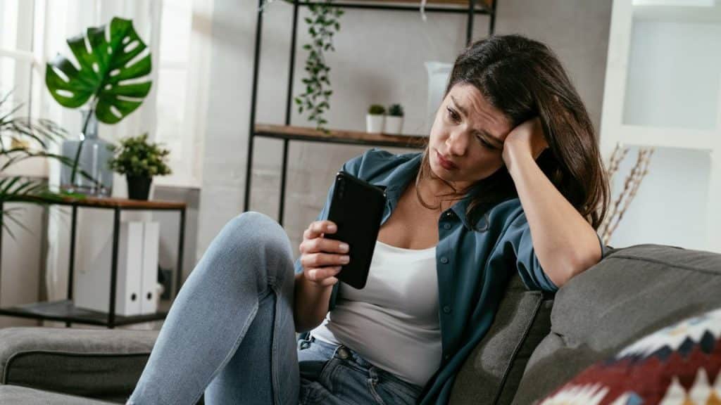 A woman sits on a couch holding a phone while resting her head on her hand.