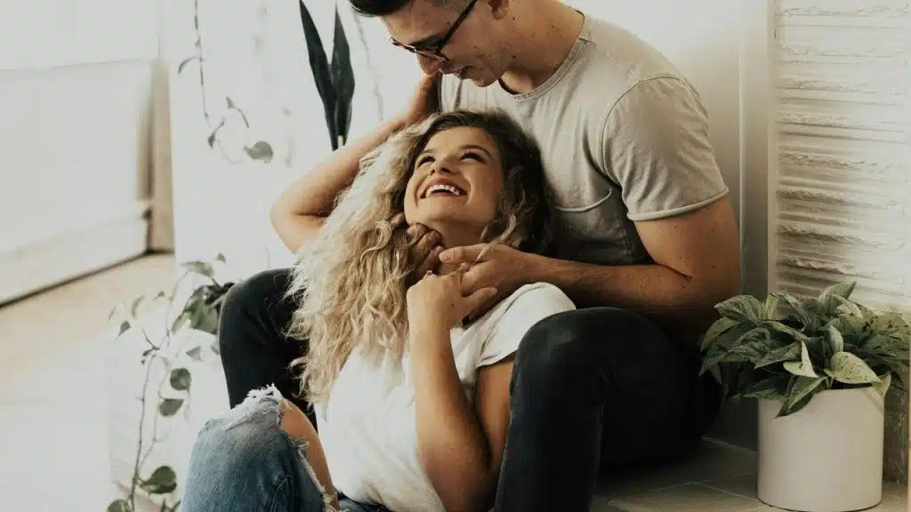 A smiling woman sitting on the floor while a man embraces her from behind.
