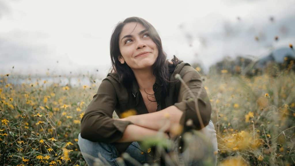 A woman sitting in a wildflower field, looking up with a thoughtful smile.