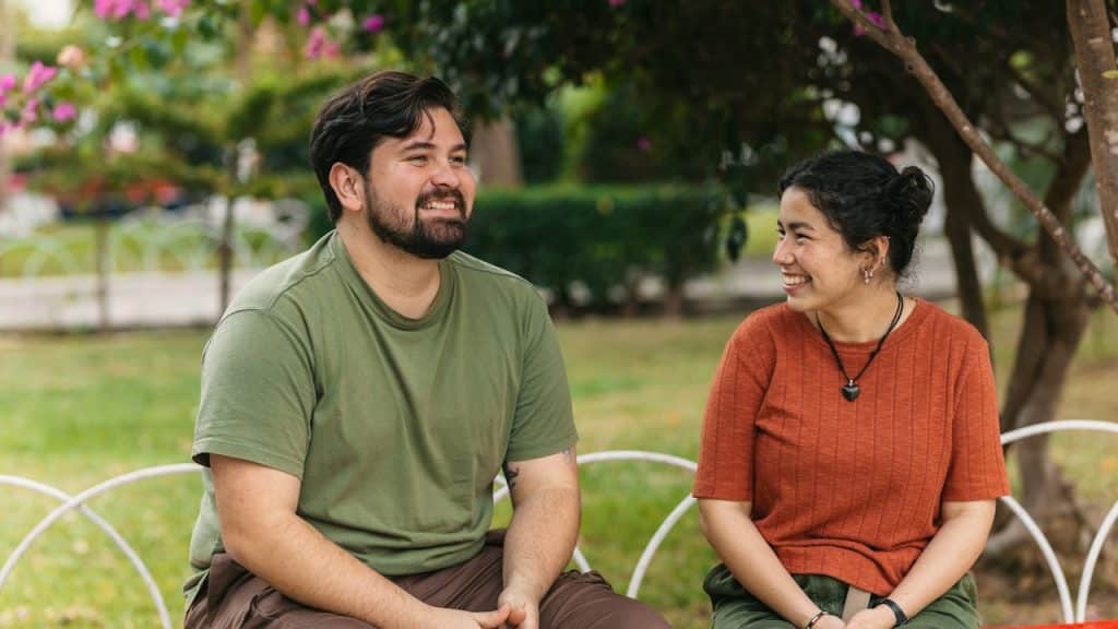 A man in a green t-shirt and a woman in an orange t-shirt sit on a bench together in a park, smiling and looking toward each other.
