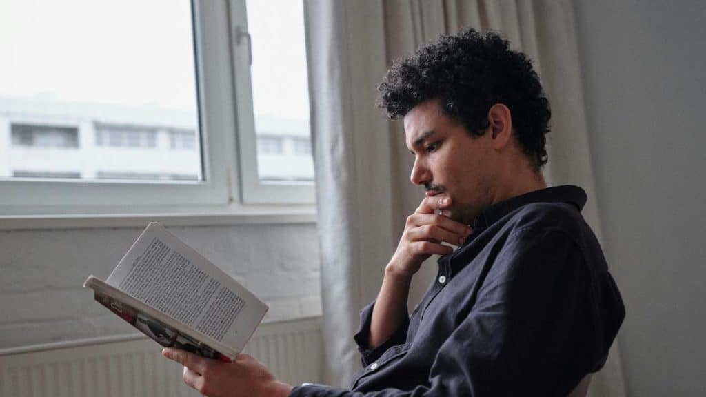 A man reading a book by a window with a thoughtful expression.