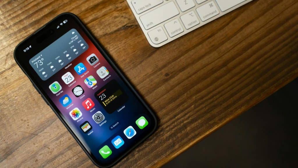 An iPhone displaying its home screen placed on a wooden desk beside a keyboard.