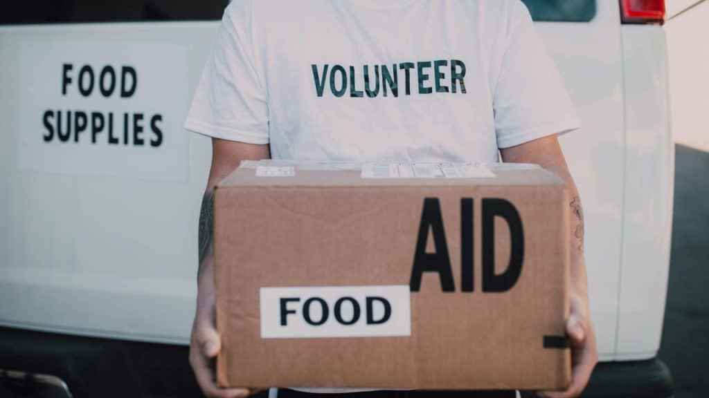 A person wearing a volunteer shirt holds a large cardboard box labeled food aid.