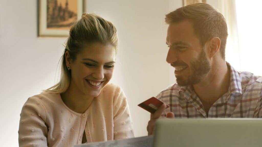 A smiling man and woman look at a laptop while the man holds a card.