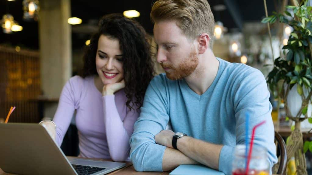 A smiling woman and a man look at a laptop screen together in a cafe.