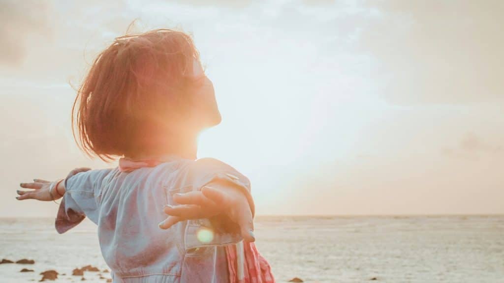 A woman standing with her hands outstretched by the ocean while the sun shines in the background.