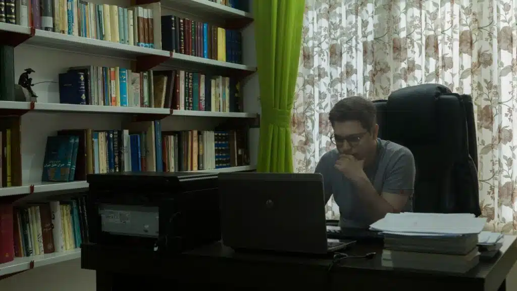 A man with glasses sits at a desk, looking intently at his laptop.