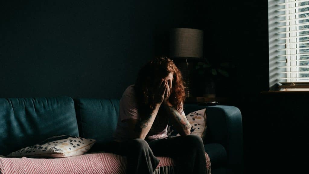A woman sitting on a couch in a dimly lit room, head in hands, appearing distressed, with light coming through window blinds.
