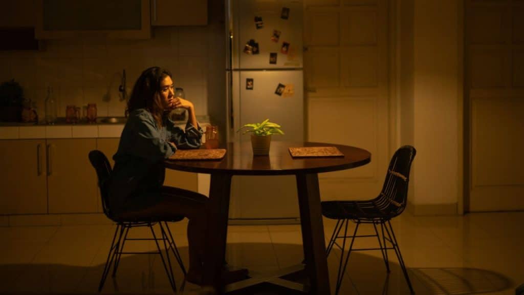 A woman sits alone at a circular wooden table in a dimly lit kitchen area.
