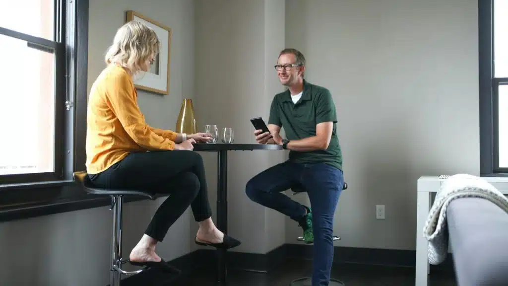 A couple sitting at a desk and talking about something while the bespectacled man holds his phone.