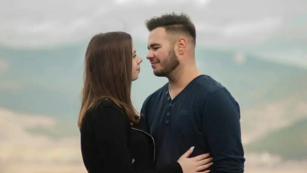 A man and woman stand close together outdoors, looking into each other's eyes.