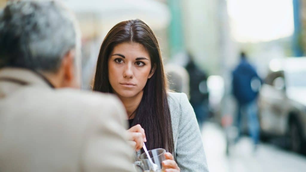 A woman with long dark hair stares forward while sitting at a table with someone.