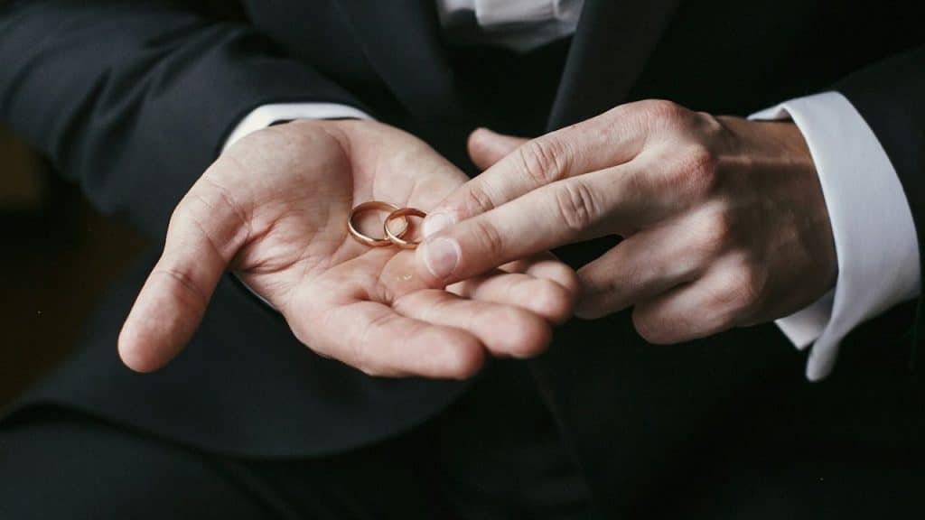 A person wearing a dark suit holds two gold wedding bands in the palm of their hand.