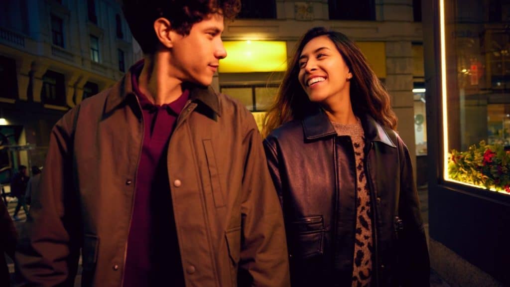 A man and woman walk together at night, smiling and looking at each other.