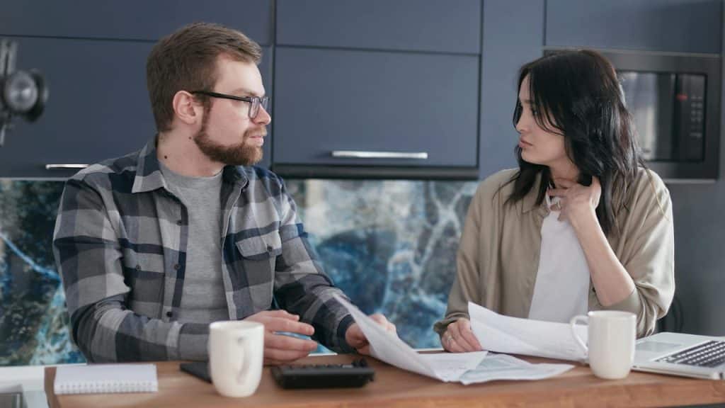 A man and woman sit at a counter with papers, a calculator, and two mugs.